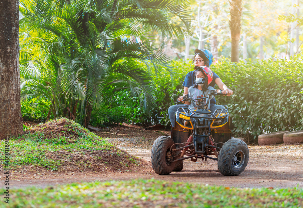 Fototapeta premium Asian mother and daughter riding quad bike in farm of Thailand, travel while covid-19, child has medical mask on face, medical mask with strap hang on neck. Background of green trees in farm.