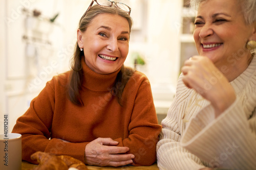 Indoor image of two joyful positive European female friends on retirement having fun, talking, telling funny jokes during meeting at home or cafe. People, friendship, age and retirement concept