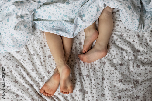 Close-up of two pairs of toddler girls feet on the bed under the blanket. Light blue and beige tones. © Светлана Густова