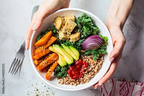 Fototapeta na wymiar Woman hands eating vegan salad of baked vegetables, avocado, tofu and buckwheat buddha bowl, top view. Plant based food concept.