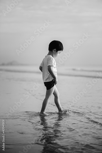 A black and white photo of a boy walking on the beach. He was looking down at his wet feet on the shallow water of the beach.