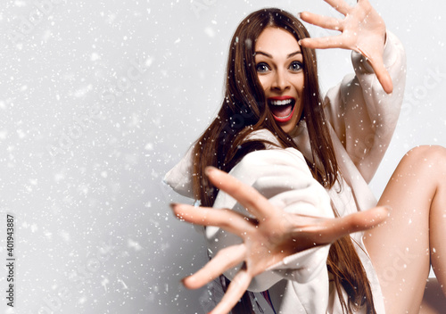 Excited woman with long hair in hoodie or sweater and naked legs in festive mood sits gesturing with hands and looking at camera under the snow over grey background, copy space. Fashion, stylish look
