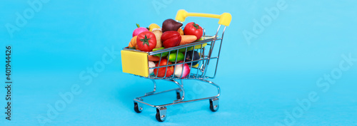 Metal shopping cart with fruits and vegetables on a blue background. Toy miniature shopping trolley