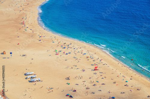 Top aerial view of sandy beach with people tourists sunbathing and Atlantic Ocean azure blue water, Praia da Nazare town coastline with waves, Leiria District, Oeste region, Portugal