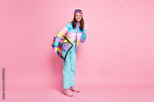 Full length photo of woman skier dressed colorful windbreaker eyeglasses holding sleigh showing v-sign isolated pink color background