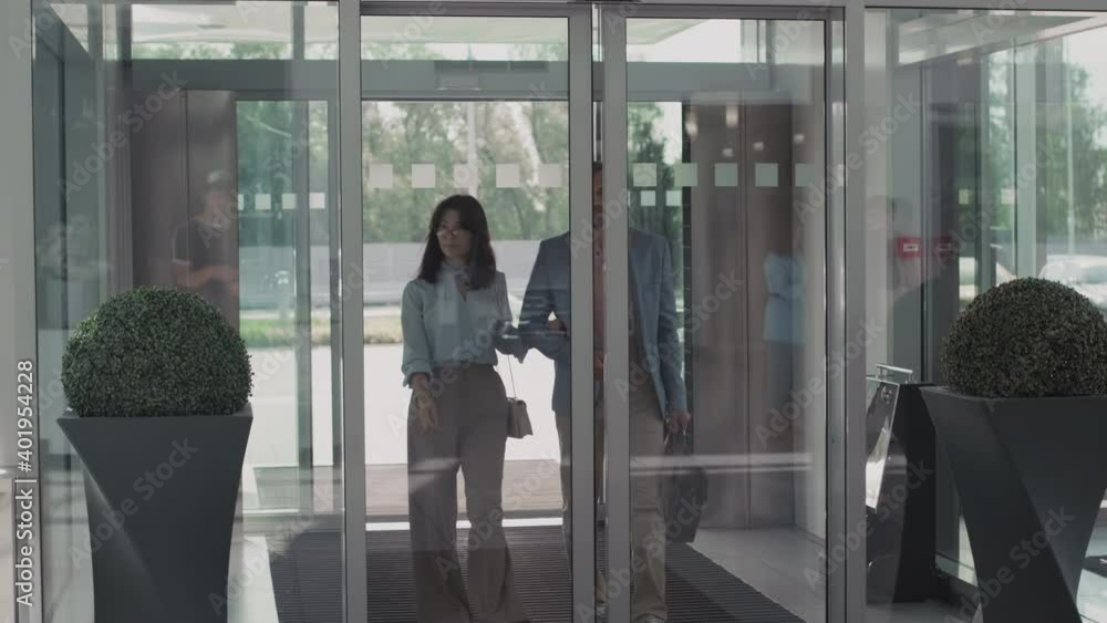 Medium shot of young woman and middle-aged man in elegant outfits walking together into car dealership through sliding glass doors Medium shot of young woman and middle-aged man in elegant outfits walking together into car dealership through sliding glass doors