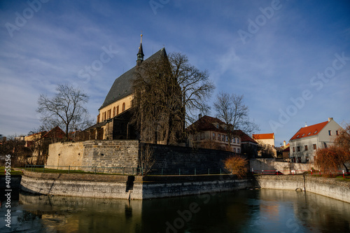 City fortifications and pond with water near gothic church of Saint Gothard with bell towers in center of medieval town Slany in sunny winter day, Central Bohemia, Czech Republic