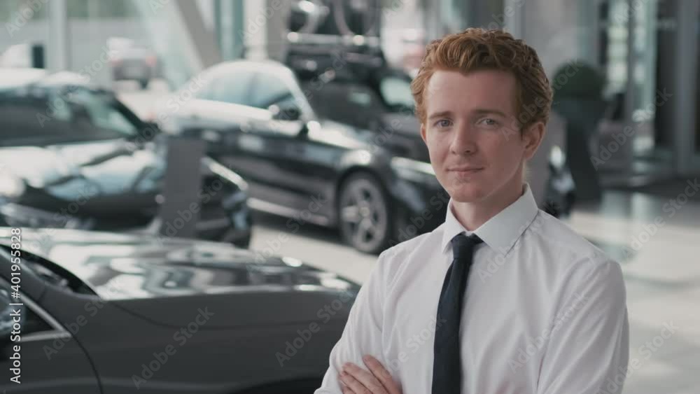 Slowmo portrait of young car salesman in necktie and white shirt standing with his arms crossed and looking at camera in dealership Slowmo portrait of young car salesman in necktie and white shirt standing with his arms crossed and looking at camera in dealership