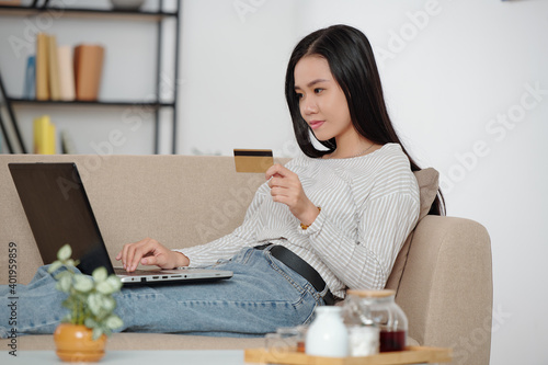 Lovely young Asian woman sitting on couch with laptop and ordering food online when staying home due to coronavirus photo