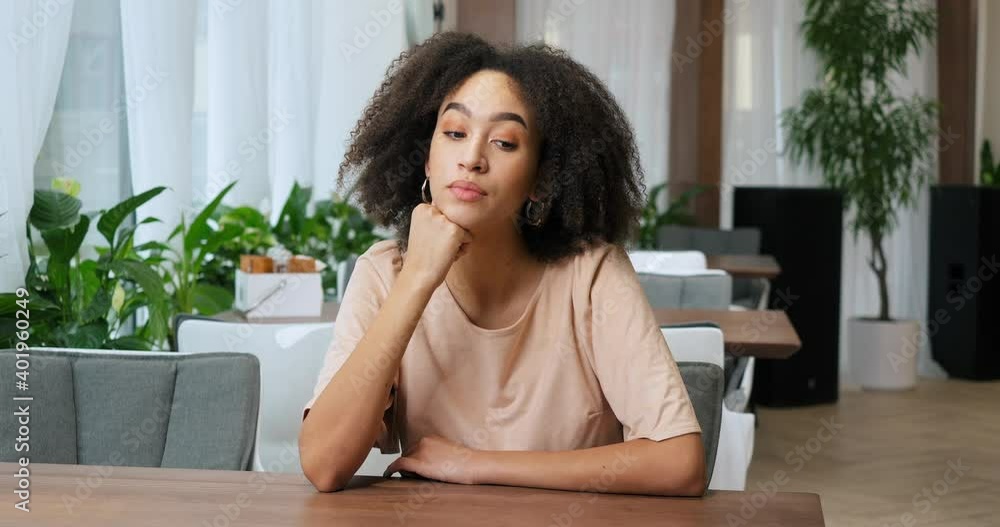 Portrait of beautiful sensual pensive millennial girl afro american woman with curly hairstyle sitting at table in cafe making choice recalls memories thinks about ideas presents future frowning face Portrait of beautiful sensual pensive millennial girl afro american woman with curly hairstyle sitting at table in cafe making choice recalls memories thinks about ideas presents future frowning face
