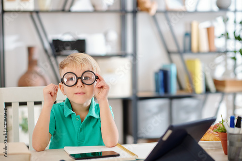Funny schoolboy putting on glasses with thick lenses and looking at camera when sitting at his desk at home