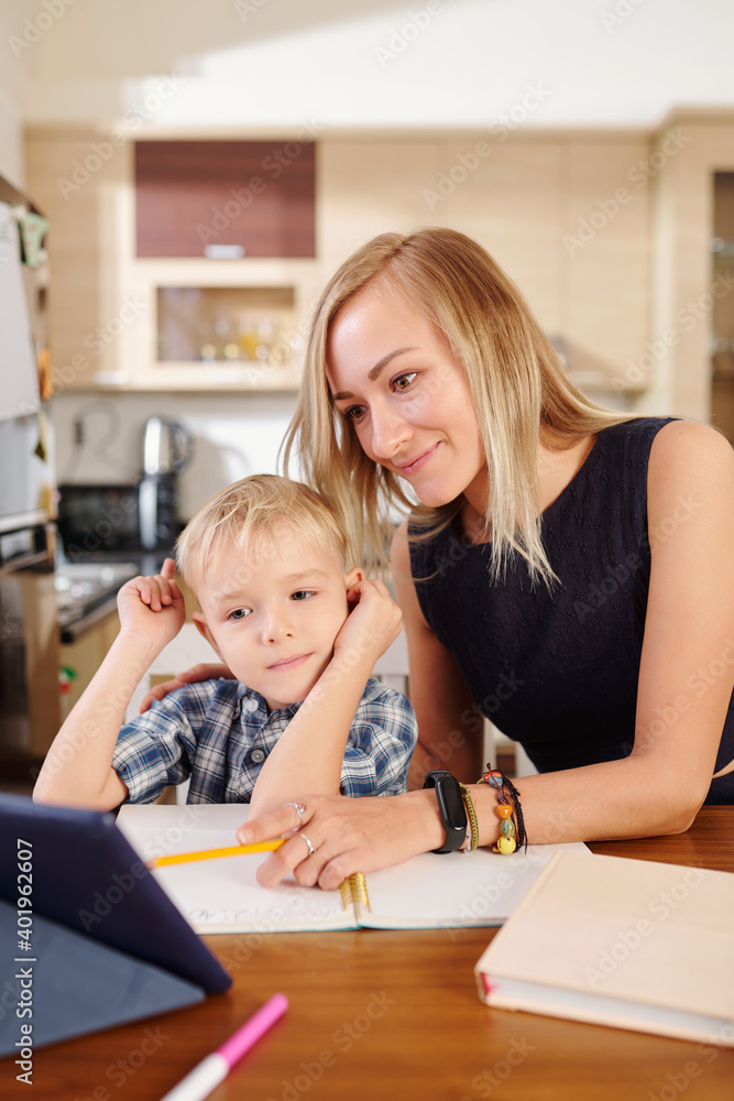 Smiling mother pointing at tablet computer when explaining lesson to her child who is studying at home due to coronavirus Smiling mother pointing at tablet computer when explaining lesson to her child who is studying at home due to coronavirus