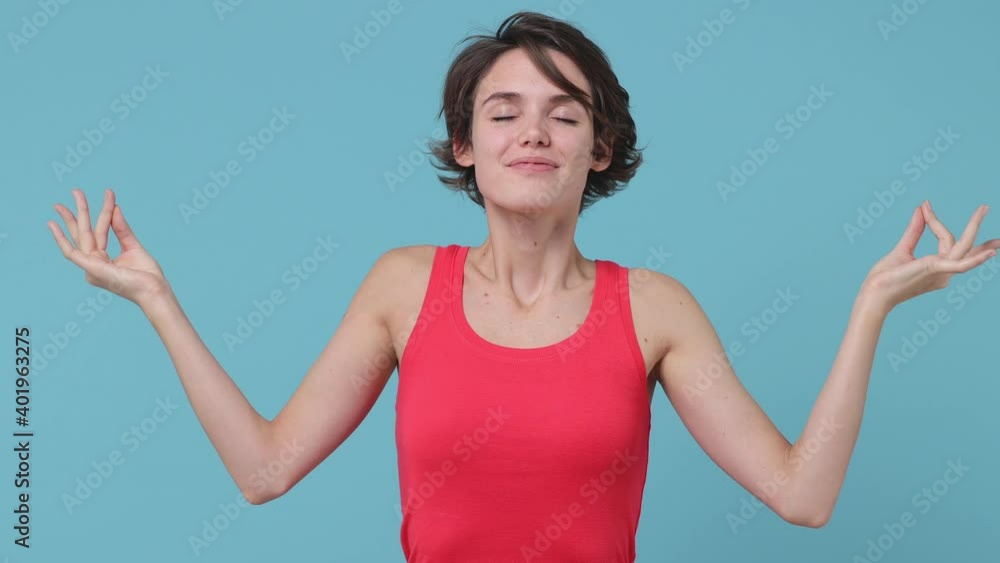 Smiling brunette young woman 20s years old in pink tank top posing isolated on blue background in studio. People lifestyle concept. Hold hands in yoga gesture relaxing meditating trying to calm down Smiling brunette young woman 20s years old in pink tank top posing isolated on blue background in studio. People lifestyle concept. Hold hands in yoga gesture relaxing meditating trying to calm down