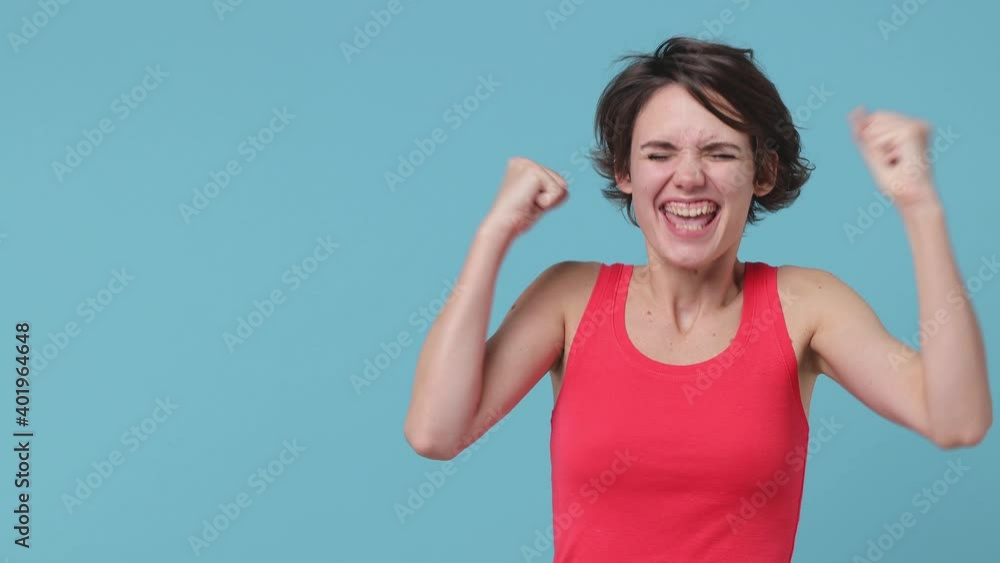 Shocked young brunette woman 20s in pink tank top posing isolated on pastel blue color background in studio. People lifestyle concept. Looking aside doing winner gesture celebrating showing thumbs up Shocked young brunette woman 20s in pink tank top posing isolated on pastel blue color background in studio. People lifestyle concept. Looking aside doing winner gesture celebrating showing thumbs up