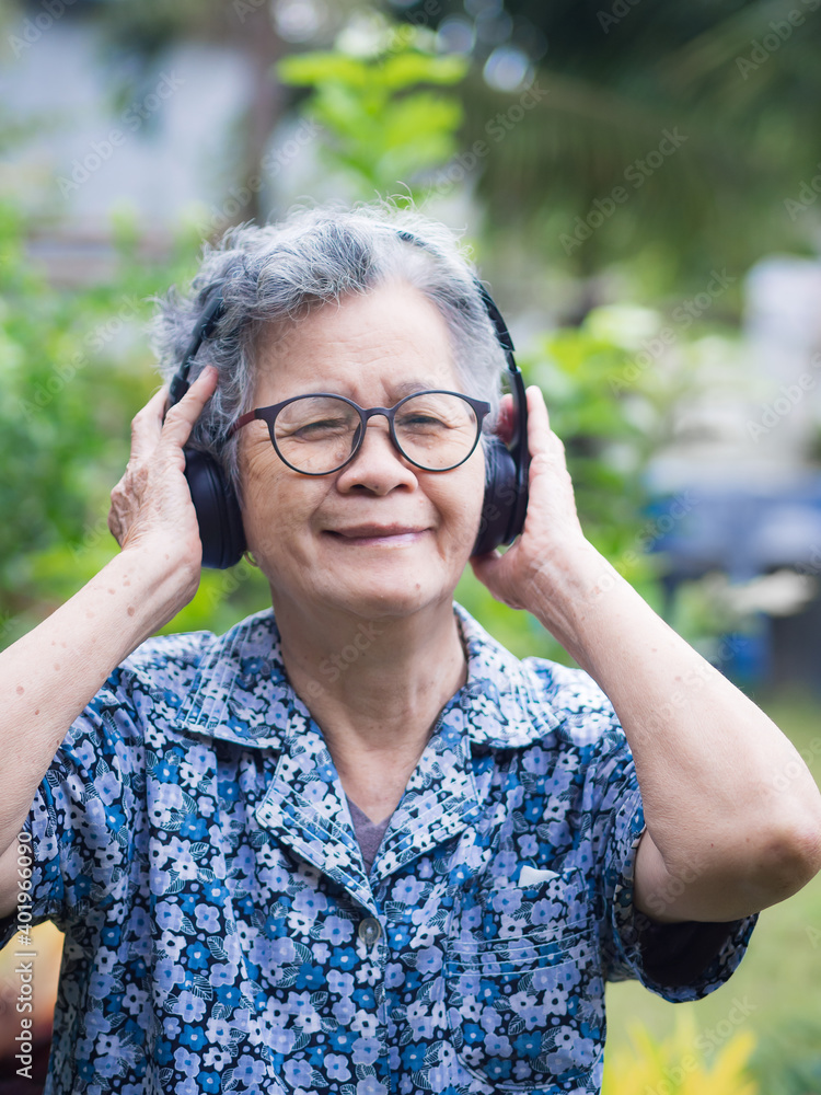A Portrait of a senior woman with short white hair wearing wireless headphones listening to a favorite song while standing in a garden. Concept of aged people and relaxation