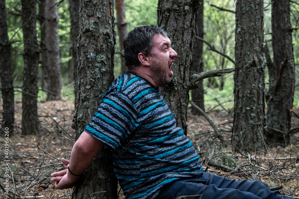 A hostage in the woods. A man in a blue t-shirt and trousers is sitting on the ground tied to a tree in the forest calling for help. Victim of an attack. Horizontal photo.