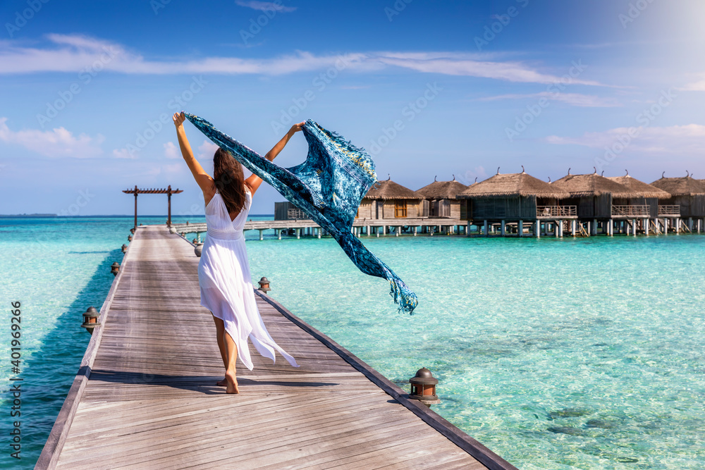 A beautiful tourist woman walks on a wooden pier over turquoise ocean in the Maldives and holds a waving scarf in her hands - obrazy, fototapety, plakaty 