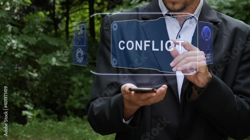 Unrecognizable businessman activates conceptual HUD holograms on smartphone with text Conflict. Bearded man in white shirt and acket with holographic screen on a background of green trees photo