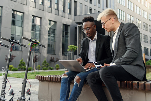 Elegant multiracial colleagues sitting on the bench near office and discussing business matters outdoors.