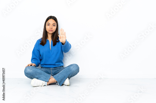 Young mixed race woman sitting on the floor isolated on white background making stop gesture