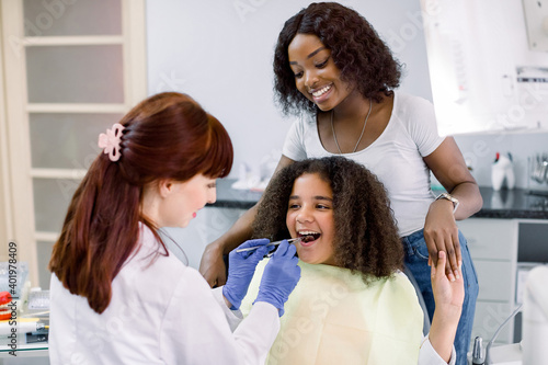 Little mixed raced school girl with curly hair, visiting dentist for checkup or caries treatment, sitting in dental chair and holding hand of her pretty African mom. Female dentist makes examination.