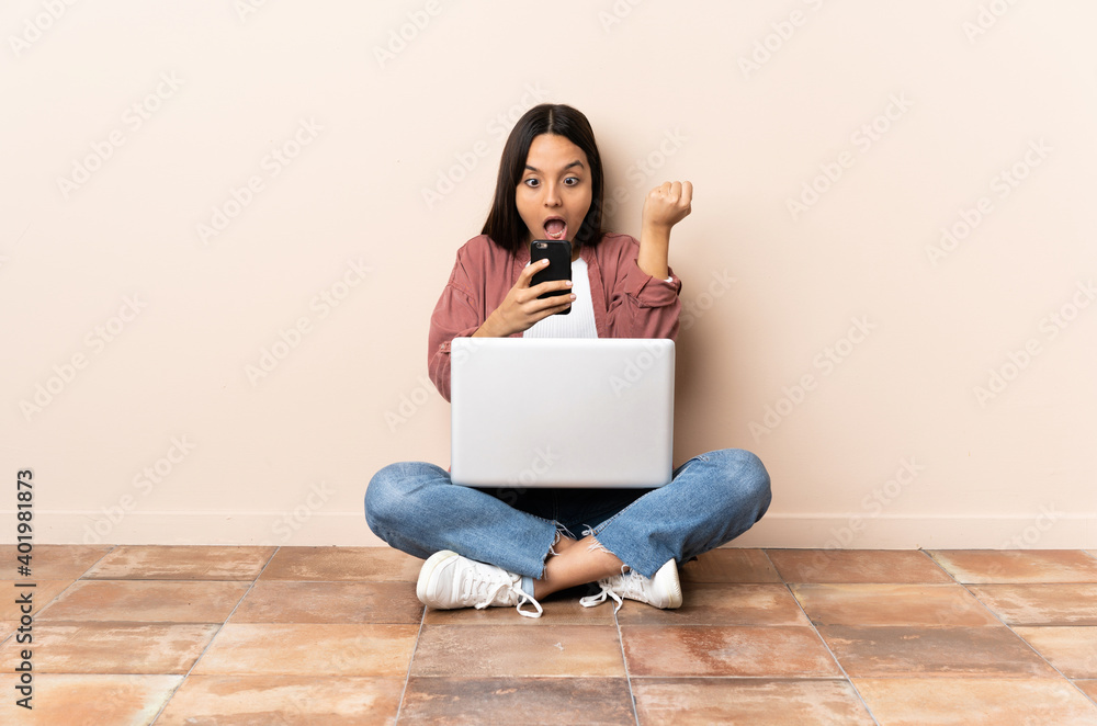 Young mixed race woman with a laptop sitting on the floor surprised and sending a message Young mixed race woman with a laptop sitting on the floor surprised and sending a message