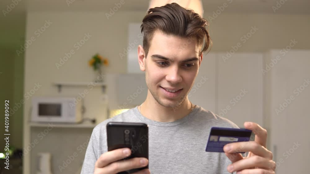 Smiling young man holding credit card and mobile phone while shopping online at home Smiling young man holding credit card and mobile phone while shopping online at home