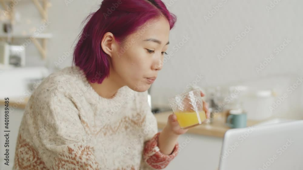 Young Asian woman with pink hair drinking orange juice and typing on laptop while having breakfast in the kitchen at home Young Asian woman with pink hair drinking orange juice and typing on laptop while having breakfast in the kitchen at home