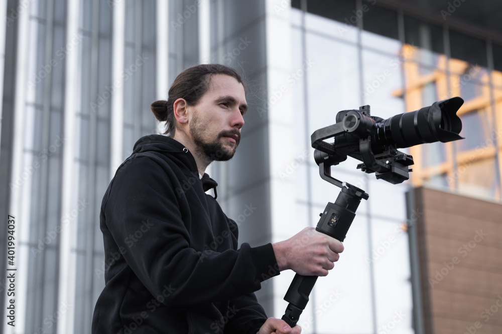 Young Professional videographer holding professional camera on 3-axis gimbal stabilizer. Pro equipment helps to make high quality video without shaking. Cameraman wearing white hoodie making a videos. Young Professional videographer holding professional camera on 3-axis gimbal stabilizer. Pro equipment helps to make high quality video without shaking. Cameraman wearing white hoodie making a videos.
