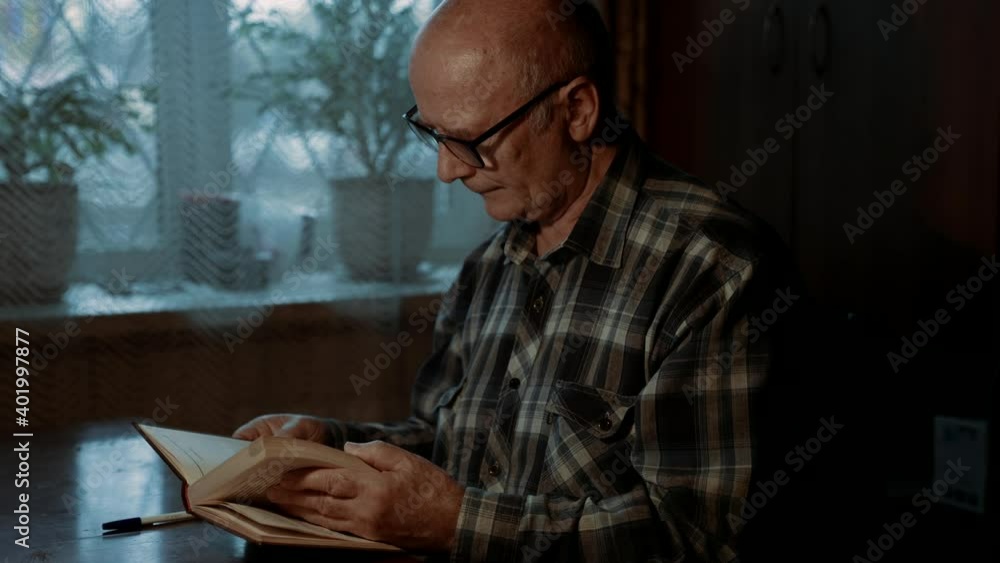 An elderly bald man with glasses attentively reads a book, sitting in a room at a table against the background of a wardrobe and a window. An elderly bald man with glasses attentively reads a book, sitting in a room at a table against the background of a wardrobe and a window.