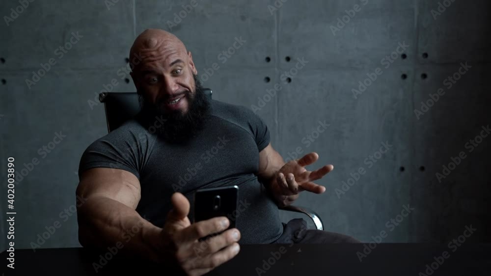 a muscular bald man sits at home at a Desk and uses a smartphone, talking on a video link, smiling and laughing. a muscular bald man sits at home at a Desk and uses a smartphone, talking on a video link, smiling and laughing.