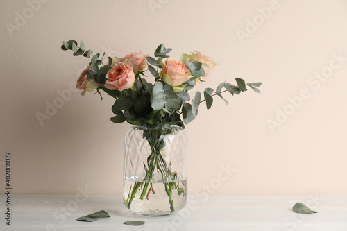 Vase with bouquet of beautiful roses on white wooden table near beige wall