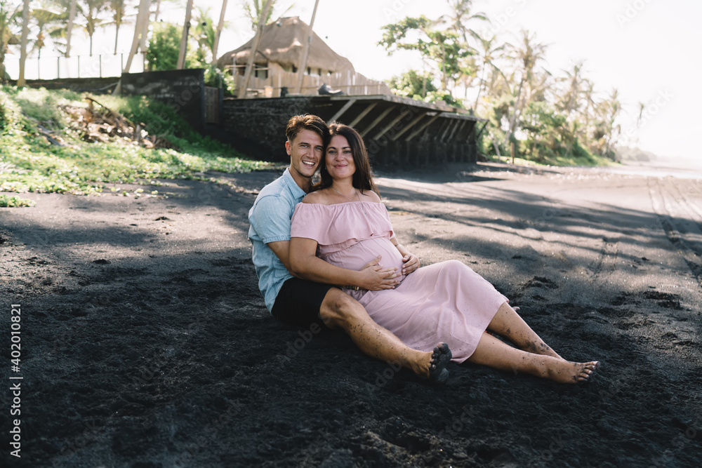Loving pregnant couple sitting on sandy beach in exotic country Loving pregnant couple sitting on sandy beach in exotic country