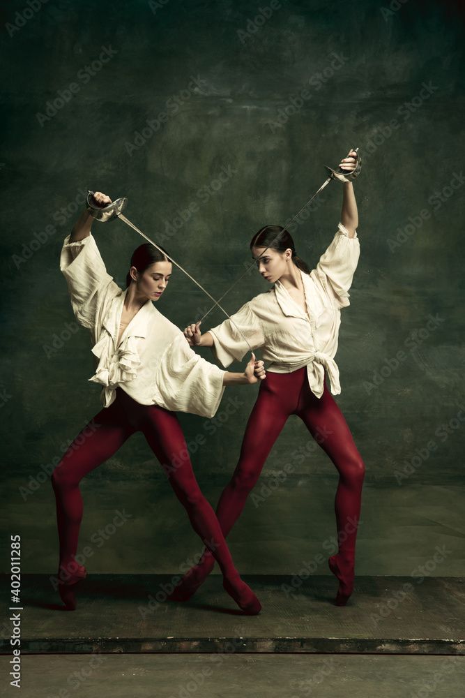 Contrast. Two young female ballet dancers like duelists with swords on dark green background. Caucasian models dancing together. Ballet and contemporary choreography concept. Creative art photo.