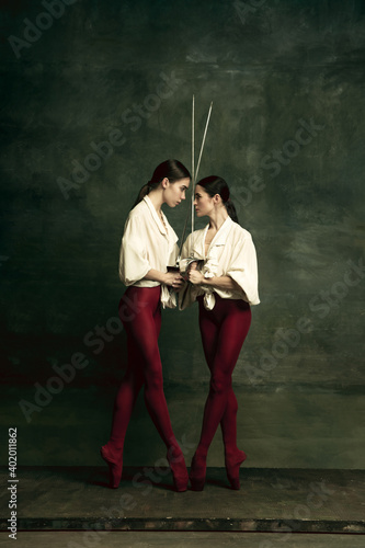 Emotions. Two young female ballet dancers like duelists with swords on dark green background. Caucasian models dancing together. Ballet and contemporary choreography concept. Creative art photo.