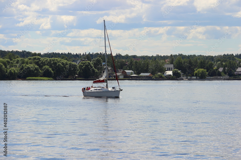 One white sailing yacht with mast without sail floats on Moscow Region water reservoir with green forest trees on far shore at summer day on blue sky with clouds background, river tourism recreation One white sailing yacht with mast without sail floats on Moscow Region water reservoir with green forest trees on far shore at summer day on blue sky with clouds background, river tourism recreation