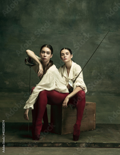 Duet. Two young female ballet dancers like duelists with swords on dark green background. Caucasian models dancing together. Ballet and contemporary choreography concept. Creative art photo.