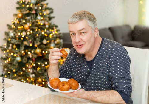Caucasian man eating olibol near the Christmas tree at home, portret,Oliebollen - Dutch traditional Dessert - Also known as Dutch Doughnuts or dutchies. photo