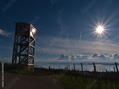 Wooden observation tower on the vineyard slopes above small village Hagnau am Bodensee, Lake Constance, Germany with bright sun in backlit reflecting on glass and blue sky in winter season.