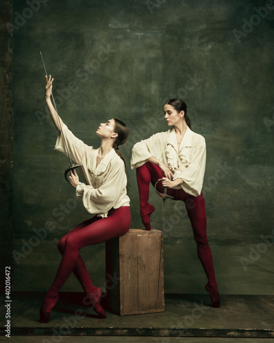 Romance. Two young female ballet dancers like duelists with swords on dark green background. Caucasian models dancing together. Ballet and contemporary choreography concept. Creative art photo.