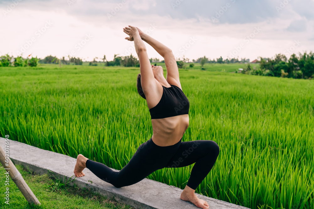 Fototapeta premium Concentrated fit girl stretching body muscles during morning pilates training near rice fields in Vietnam, calm female yogi meditating for mindfulness keeping healthy lifestyle on retreat training