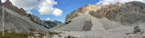 A panoramic view on a stony valley in Italian Dolomites. There are high and sharp mountains around. In the back the mountains are overgrown with green plants. Remote and raw landscape. Sunny day