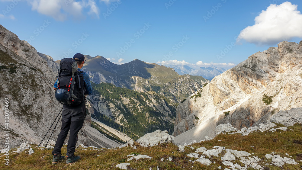 A man with a big hiking backpack admiring the view on a stony valley in Italian Dolomites. There are high and sharp mountains around. Remote and raw landscape. Sunny day. Freedom and serenity
