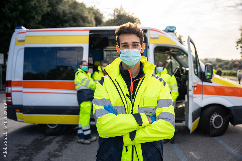 Portrait of young millennial man with arms crossed at first aid in front of his colleagues in ambulance at break time at pandemic time from Coronavirus, Covid-19 He wearing face mask and uniform work Portrait of young millennial man with arms crossed at first aid in front of his colleagues in ambulance at break time at pandemic time from Coronavirus, Covid-19 He wearing face mask and uniform work