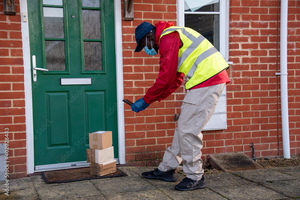Hampshire, England, UK. 2020, Male courier delivering parcels and packages during Covid-19 epidemic wearing gloves and a mask. Recording on mobile phone the delivery.