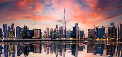 Stunning panoramic view of the Dubai skyline at sunset with buildings and skyscrapers reflected on a silky smooth water flowing in the foreground. Dubai, United Arab Emirates.