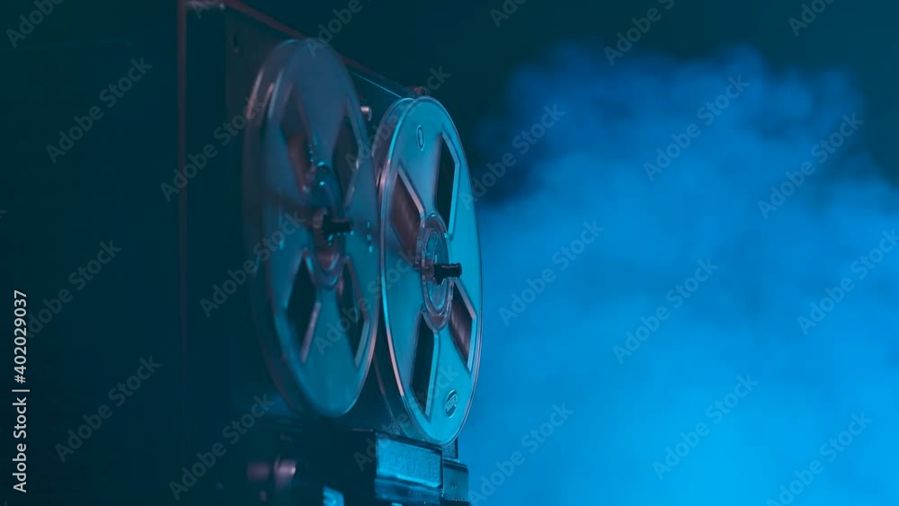 Black reel to reel tape recorder playing against a smoky studio background with blue neon lights. Rewinds retro tape. Vintage music player. Spinning reels transparent close up. Slow motion.