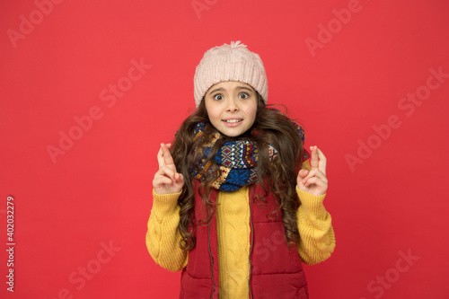 surprised pretty kid girl in puffer waistcoat with knitted hat and sweater hold crossed fingers, make a wish