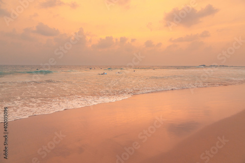 Ocean beach under beautiful sunset sky with clouds and its reflections in the wet sand of a coast. Surfers floating on waves in expectation.