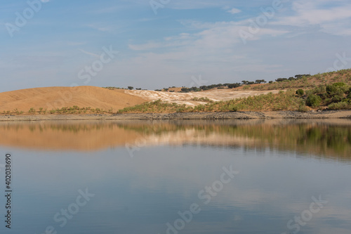 Desert like hill landscape with reflection on the water on a dam lake reservoir with blue sky in Terena, Portugal